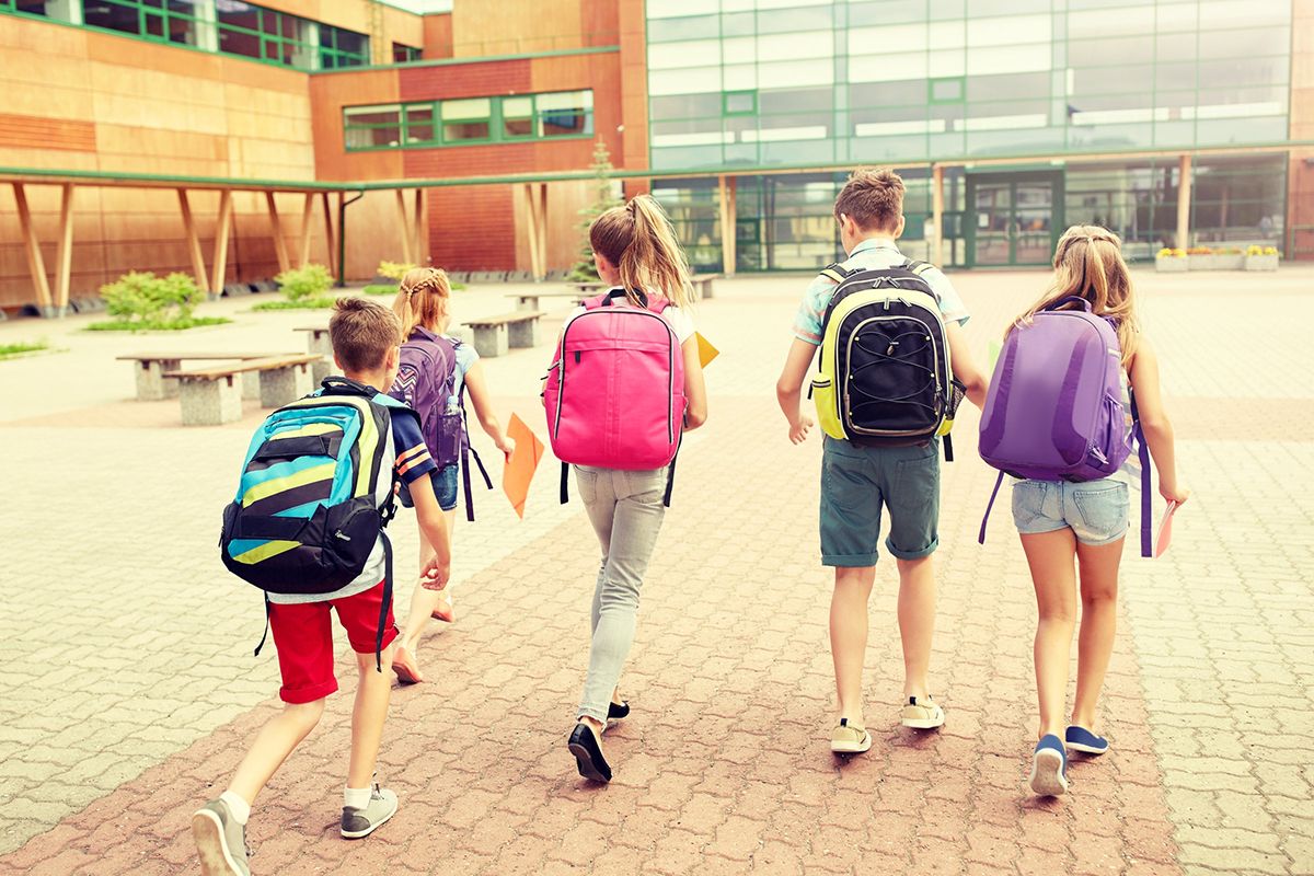 Un grupo de 5 niños de espaldas a la escuela. Cada niño lleva una mochila o bolsa escolar de color