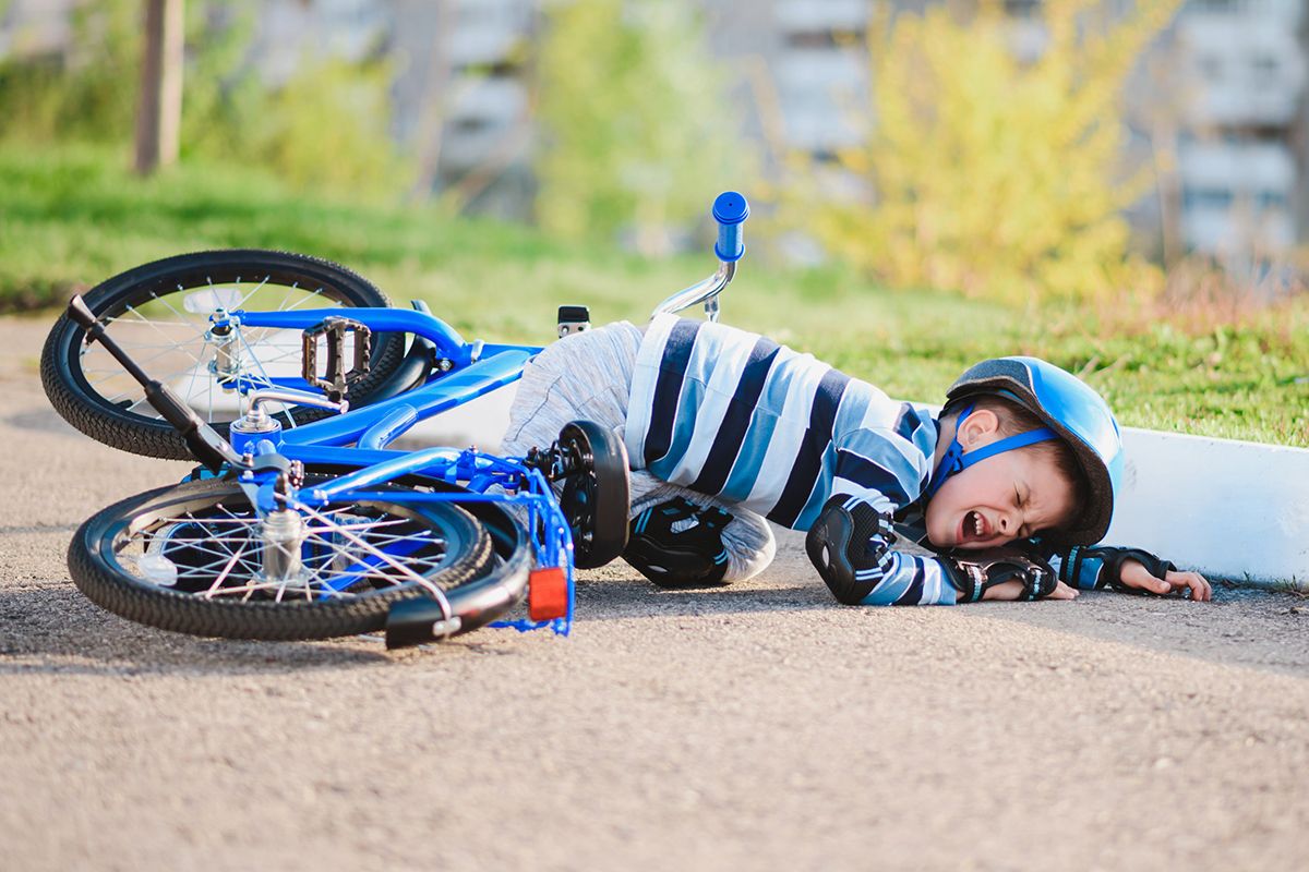 Un niño pequeño que se ha caído de su bicicleta azul. Está llorando y lleva casco de ciclista, rodilleras y guantes protectores negros