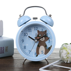 Reloj despertador infantil azul con motivo de mapache marrón, colocado sobre un mueble marrón junto a una flor blanca