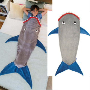 La foto está dividida en dos partes, la primera muestra a un niño tumbado en la cama, metido en un saco de dormir gris con forma de tiburón, aletas azules y boca roja. La segunda parte de la imagen muestra este saco de dormir sobre un fondo blanco.